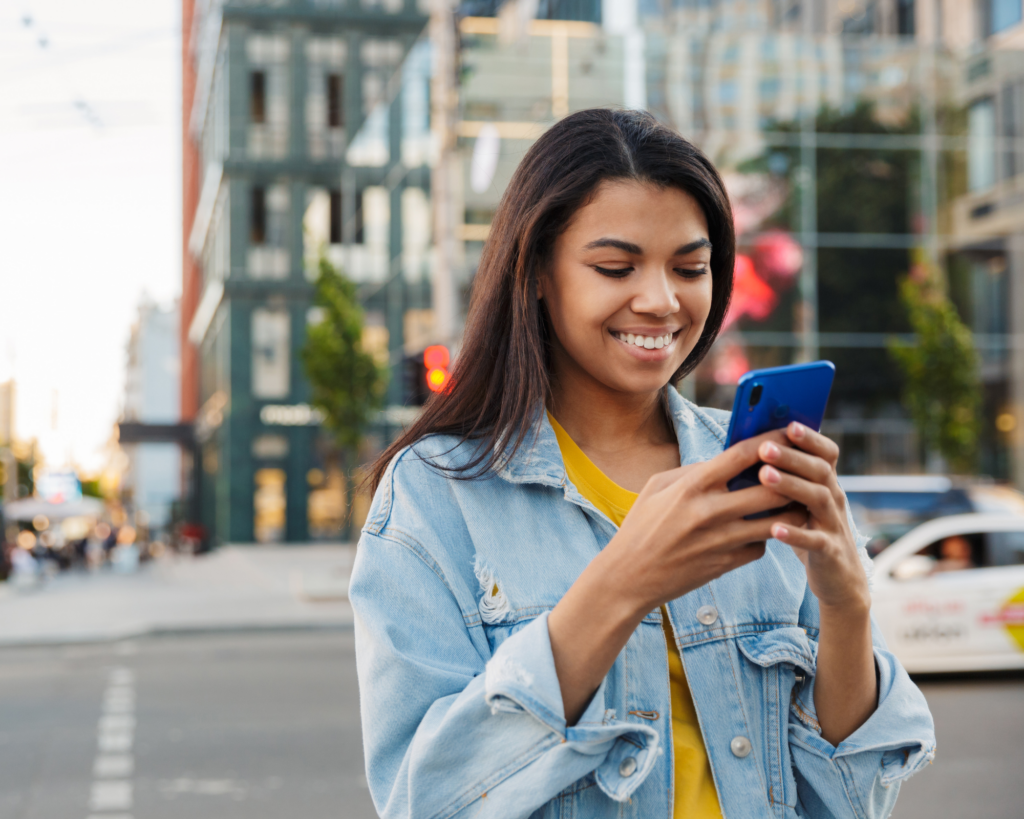 Mujer joven sonriente usando un teléfono móvil en una ciudad moderna. Lleva una chaqueta de mezclilla y una camiseta amarilla mientras se encuentra en una calle concurrida con edificios y automóviles de fondo.