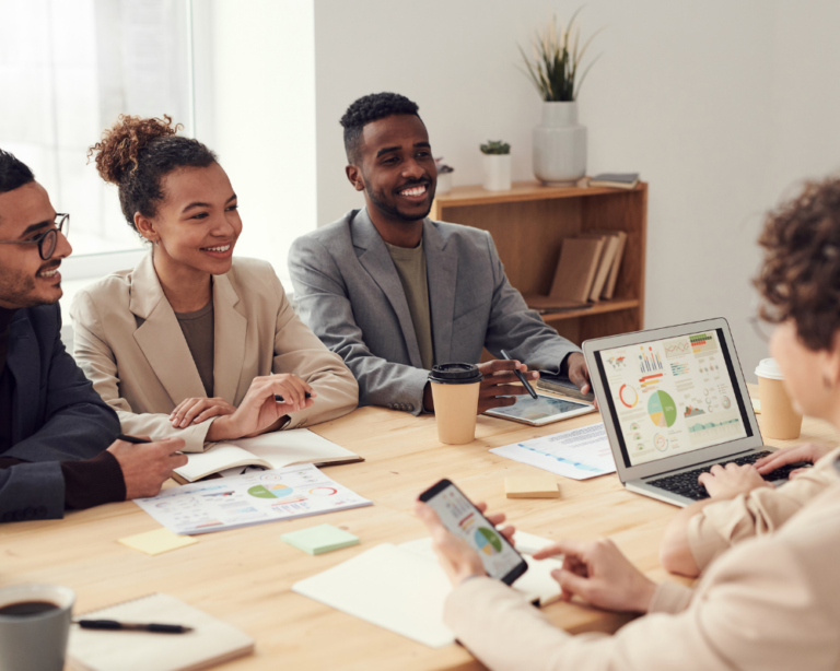 La imagen muestra a un grupo diverso de profesionales en una reunión de trabajo, sentados alrededor de una mesa en una oficina bien iluminada. Cuatro personas (dos hombres y dos mujeres) participan activamente en la discusión, sonriendo y mirando hacia un portátil que muestra gráficos coloridos y datos. Todos están vestidos con ropa de negocios casual. El ambiente es colaborativo y positivo, reflejando un entorno de trabajo moderno y dinámico. A la derecha, se puede ver una mesa auxiliar con plantas, lo que añade un toque de frescura y vida al espacio de trabajo.